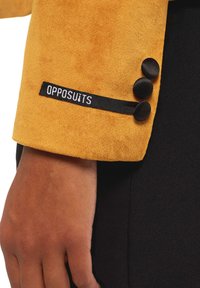 Manga de blazer de ante amarillo con tres botones negros y una etiqueta de OPPOSUITS. Mano y parte de tela negra visible.