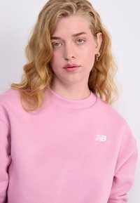 Sweatshirt rose clair en tissu doux, avec un col côtelé et un petit logo blanc sur la poitrine gauche. Texture lisse.