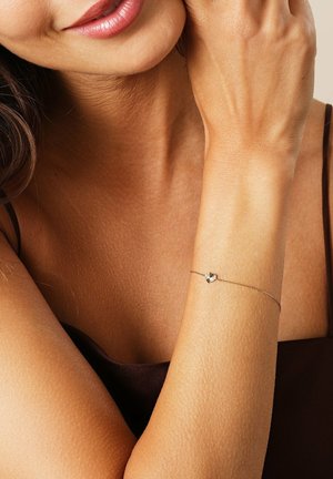 Guldtonet armbånd med en struktureret hjertecharme, der har en delikat kæde og subtil glans, båret på en solbrun arm.