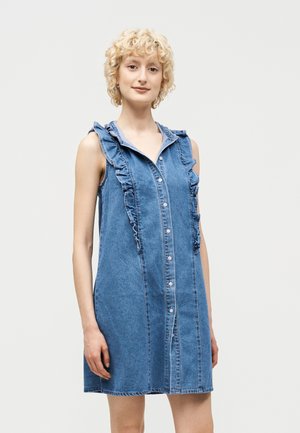 Jonge vrouw met kort, krullend blond haar, gekleed in een mouwloze blauwe spijkerjurk met ruches en knopen aan de voorkant, tegen een effen lichte achtergrond.