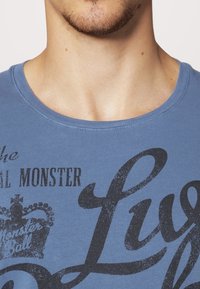 Modré bavlněné tričko s kulatým výstřihem, na kterém je velký černý grafický potisk zahrnující text a design koruny. Hladká textura.