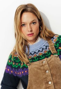 Pull en tricot avec un empiècement bleu et à motifs, présentant des designs verts, violets et orange ; associé à des salopettes en velours côtelé marron avec des boutons.