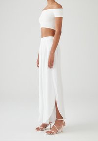 Witte off-the-shoulder crop top en lange plisségordijn, beide gemaakt van lichte stof. Bevat open split en strappy witte sandalen.