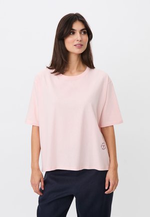 Młoda kobieta z długimi ciemnymi włosami, ubrana w luźny blady różowy t-shirt i ciemne spodnie, patrząca lekko na bok na jednolitym tle.