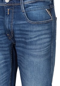 Gros plan sur un jean en denim bleu montrant une poche avant, une fermeture par bouton et une texture délavée avec des détails de coutures orange.