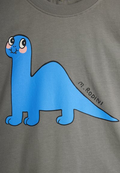 Dinosaur bleu en cartoon avec des joues roses et de grands yeux sur un tissu gris, avec le texte "M. Rodini" à côté de la queue.