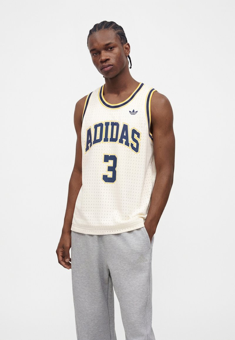 Young man with braided hair wearing white Adidas sleeveless jersey with number 3 and light gray Adidas sweatpants standing against white background.