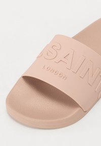 Světle růžové skluzavkové sandály vyrobené z texturované gumy, se širokým páskem embosovaným s nápisem "SAINT LONDON" pro branding. Plochý design podešve.