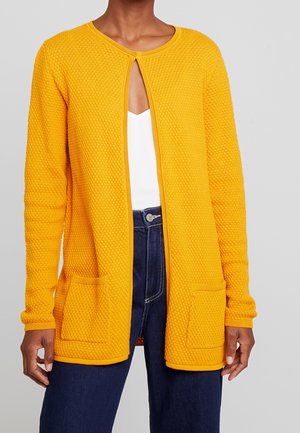 Vrouw die een gestructureerde, felgele openvallende cardigan met zakken draagt, een wit shirt en donkerblauwe spijkerbroek, tegen een effen achtergrond.