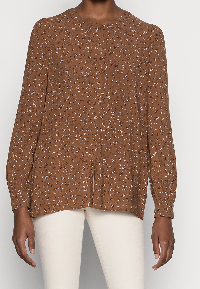 Blusa floreale marrone con maniche lunghe, scollo rotondo, chiusura con bottoni e vestibilità ampia, decorata con motivi floreali bianchi e blu.