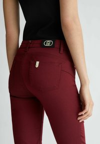 Jeansy dżinsowe w kolorze bordowym o wąskim kroju, z czarną metką z logo, metalowymi elementami oraz zdobieniami na tylnych kieszeniach. Gładka tekstura.