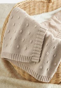 Coperta in maglia beige chiaro con motivi a pallini testurizzati, bordi a costine e una trama morbida, poggiata su un cesto intrecciato.