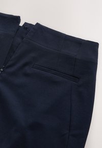 Pantalon bleu marine en tissu lisse avec une fermeture éclair et une poche arrière ; présente des lignes épurées et une coupe ajustée.