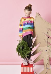 Suéter de punto colorido con rayas horizontales en rosa, naranja, amarillo, verde y azul. Pantalones oscuros y una corona verde en la mano; árbol de cartón detrás.