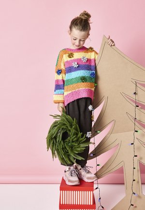 Suéter de punto colorido con rayas horizontales en rosa, naranja, amarillo, verde y azul. Pantalones oscuros y una corona verde en la mano; árbol de cartón detrás.