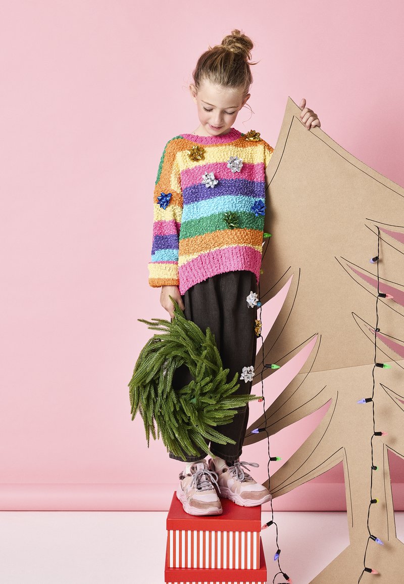 Suéter de punto colorido con rayas horizontales en rosa, naranja, amarillo, verde y azul. Pantalones oscuros y una corona verde en la mano; árbol de cartón detrás.