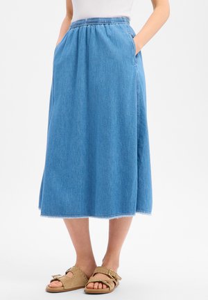 Frau trägt einen midilangen blauen Jeansrock mit elastischem Bund und Seitentaschen, kombiniert mit beigen Sandalen mit Nieten.