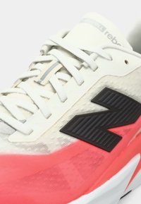 Sportlicher Schuh mit einem hellgrauen Mesh-Obermaterial, einem schwarzen "N"-Logo, rosa Akzenten an der Sohle und weißen Schnürsenkeln. Strukturierte Oberflächendetails sichtbar.