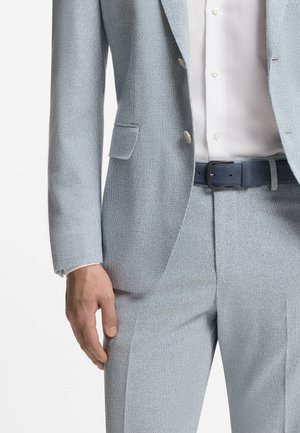 Uomo che indossa un abito grigio chiaro strutturato con blazer e pantaloni abbinati, camicia bianca abbottonata e cintura blu scuro, mano rilassata al lato.