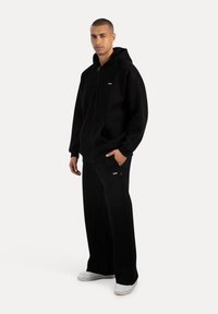 Sort zip-up hættetrøje med en kængurulomme og et lille logo, sammen med brede sorte bukser og hvide sneakers på en neutral baggrund.