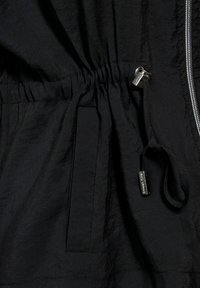 Svart lättviktjacka med elastisk dragsko i midjan, dragkedjestängning och en sidoficka. Har en slät textur och metalliska detaljer.