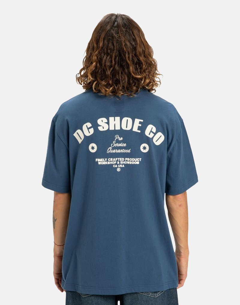Person med axellångt lockigt hår som bär en blå T-shirt från DC Shoe Co. med vit text på ryggen, står mot en enfärgad ljus bakgrund.