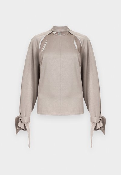 Blouse beige à manches longues avec épaules découpées, col haut et poignets noués, présentée sur un fond clair uni.