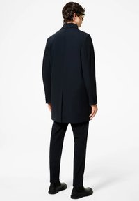 Cappotto blu scuro con colletto, fessura singola e maniche abbottonate, abbinato a pantaloni neri con texture e scarpe nere. Design semplice e moderno.