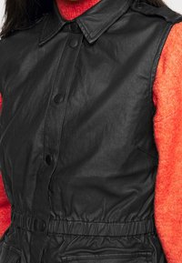 Musta nahkatakki, jossa on kaulus, painonapit ja joustava vyötärönauha. Sileä pinta ja sivutaskut. Päällä punaisen neuleen päällä.