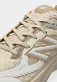 Baskets New Balance beige et crème avec dessus en mesh, lacets épais et logo "N" proéminent sur le côté.