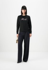 Femme portant un sweat-shirt noir avec le texte "Karl", un large pantalon marine, des chaussures noires, tenant un sac à main beige, se tenant contre un fond blanc.