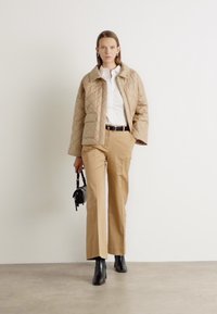 Beige quilted jacket with a collar, white shirt underneath, and beige trousers. Black ankle boots and a small black handbag complete the outfit.
