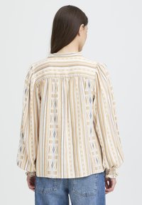 Blouse à manches longues en tissu à rayures jaunes et blanches avec des motifs géométriques. Présente des épaules froncées et un ourlet arrondi.