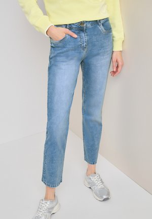 Person iført lyseblå denimjeans, en gul sweatshirt og sølvhvide sneakers, stående med den ene hånd i lommen.