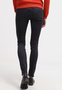 Vaqueros de mezclilla negros de corte ajustado, con detalles de costura en los bolsillos traseros, combinados con un suéter naranja texturizado y zapatos negros.