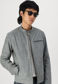 Jachetă din faux suede de culoare gri deschis, cu guler în picioare, buzunar pe piept cu fermoar și două buzunare laterale. Modelul poartă ochelari de soare negri, punând în evidență un design minimalist.