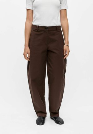 Personne portant un pantalon marron ample, une chemise blanche à manches courtes, des chaussures noires et un bracelet argenté au poignet droit.