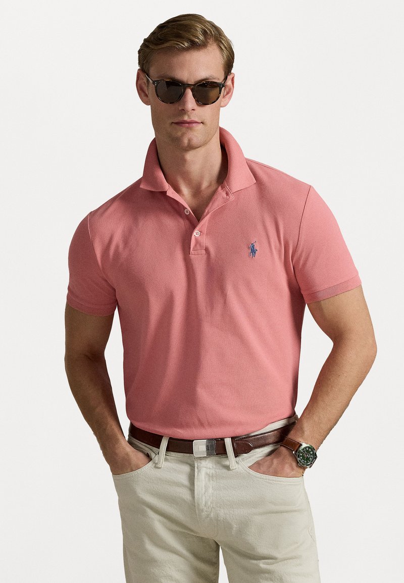 Cămașă polo roz din bumbac texturat cu guler ribbed, având un logo albastru. Asortată cu pantaloni bej și o curea maronie, accesorizată cu ochelari de soare.