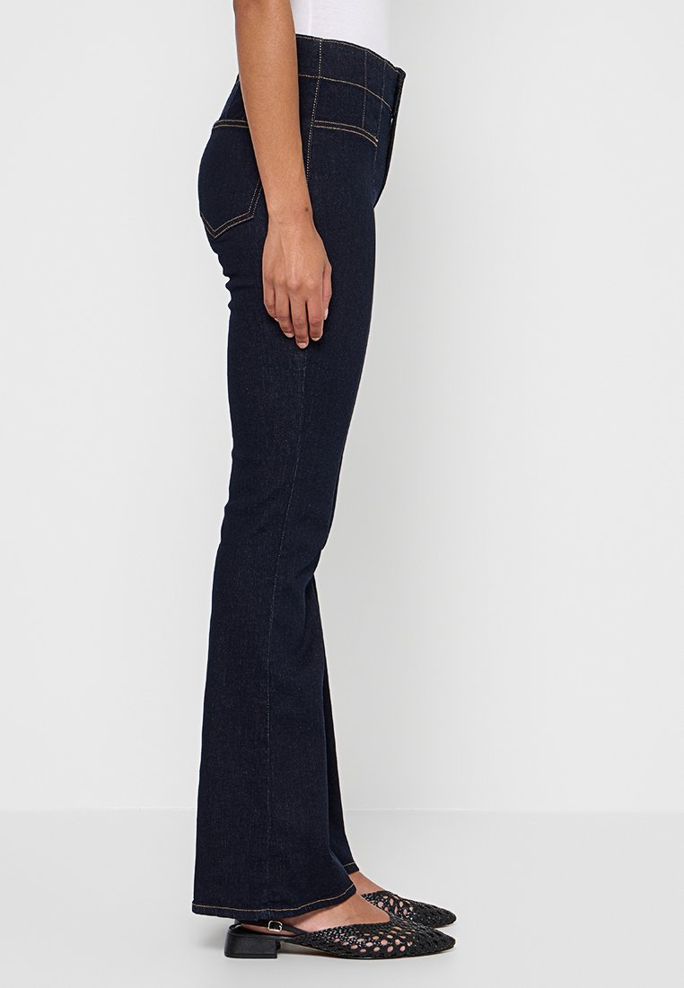 Jean évasé bleu foncé en denim, avec des coutures dorées, une taille mi-haute et une silhouette ajustée qui s'évase aux chevilles.