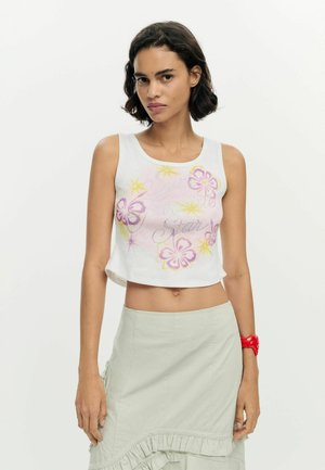 Jeune femme aux cheveux courts et foncés portant un haut court blanc à fleurs sans manches et une jupe beige clair avec des détails à volants.