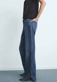 Vaqueros de denim azul de cintura alta y pierna ancha con aberturas laterales en el dobladillo; combinados con una blusa negra. Textura suave, diseño clásico de cinco bolsillos.