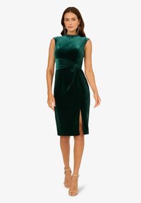 Zielona aksamitna sukienka o dopasowanym kroju, z wysokim dekoltem, rozporkiem po boku i marszczeniem w talii. Noszona z nagimi sandałami na obcasie.
