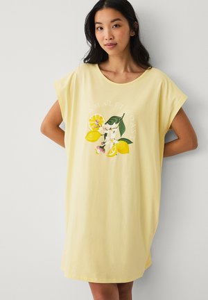 Kvinde iført en løs gul kjole med print af citroner og blomster samt teksten "Amalfi Coast" på forsiden, stående op ad en hvid væg.