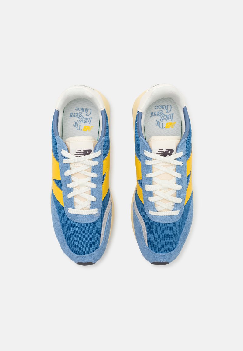 Un par de zapatillas New Balance en azul y amarillo con cordones blancos y el logo visible en la lengua, vistas desde arriba sobre un fondo blanco.
