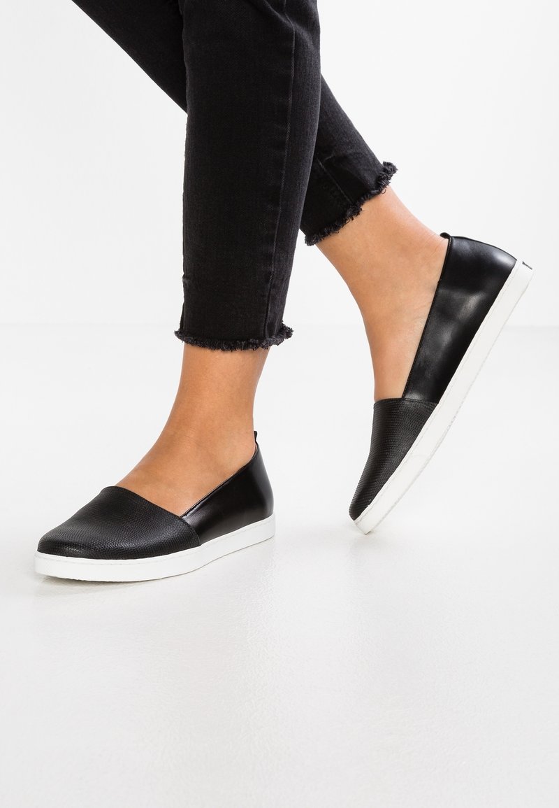 Pieds portant des chaussures noires à enfiler avec des semelles blanches, associées à un jean noir raccourci sur un fond blanc uni.
