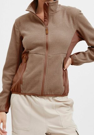 Femme portant une veste marron texturée zippée avec poches latérales et pantalon crème taille haute, main sur la hanche, sur fond uni.
