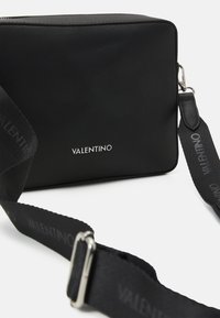 Borsa rettangolare nera realizzata in materiale testurizzato, con logo argentato, tracolla regolabile e dettagli a contrasto sulla tracolla.