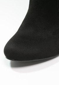 Svart mocka ankelboots med spetsig tå och slät yta; har en låg, platt gummisula. Närbild betonar materialdetaljer.