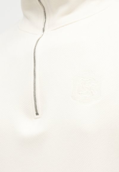 Ενδυμα από λευκό υφασμάτινο ύφασμα με υφή, ασημένιο μισό φερμουάρ και διακριτικό ανάγλυφο έμβλημα στο στήθος.