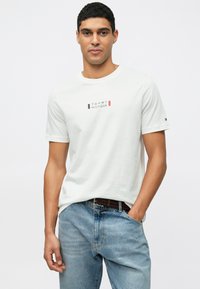 Λευκή βαμβακερή μπλούζα τύπου T-shirt με στρογγυλή λαιμόκοψη, που διαθέτει τυπωμένο το λογότυπο "TOMMY HILFIGER" σε κόκκινο και μπλε χρώμα. Συνδυάζεται με μπλε τζιν παντελόνι.